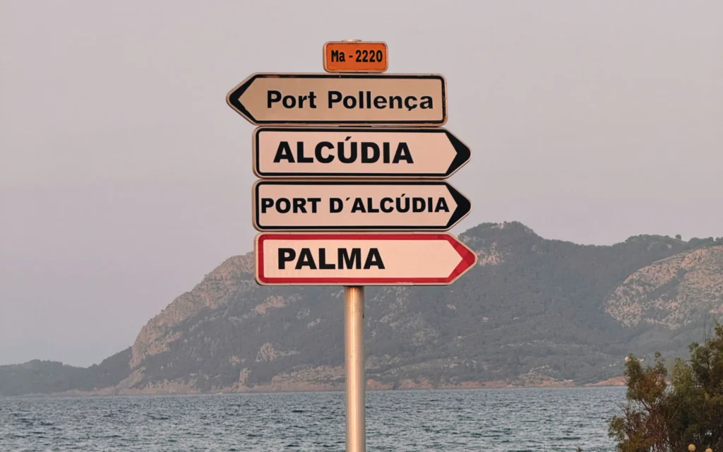Señales hacia Port Pollença, Alcúdia y Palma con mar y montaña al fondo.
