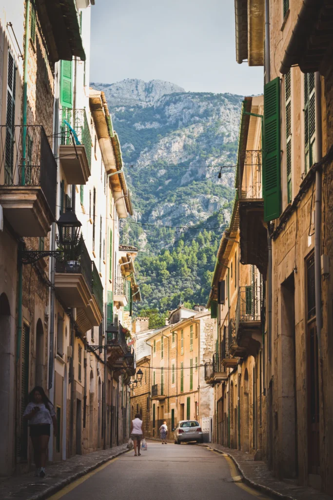 Calle entre casas de piedra con persianas verdes y una montaña al fondo.